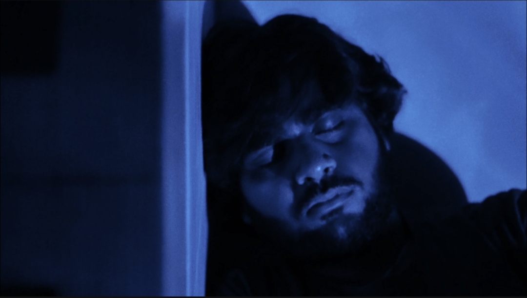 A blue-lit close-up of the protagonist resting against a wall in Qaid.