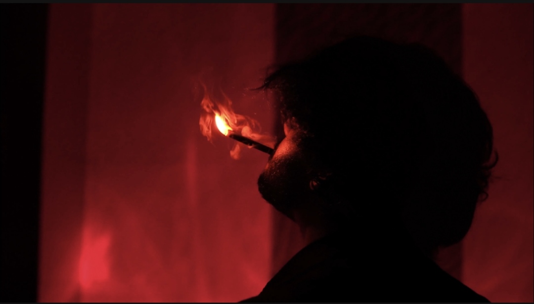 The protagonist lighting a cigarette against a red background in Qaid.